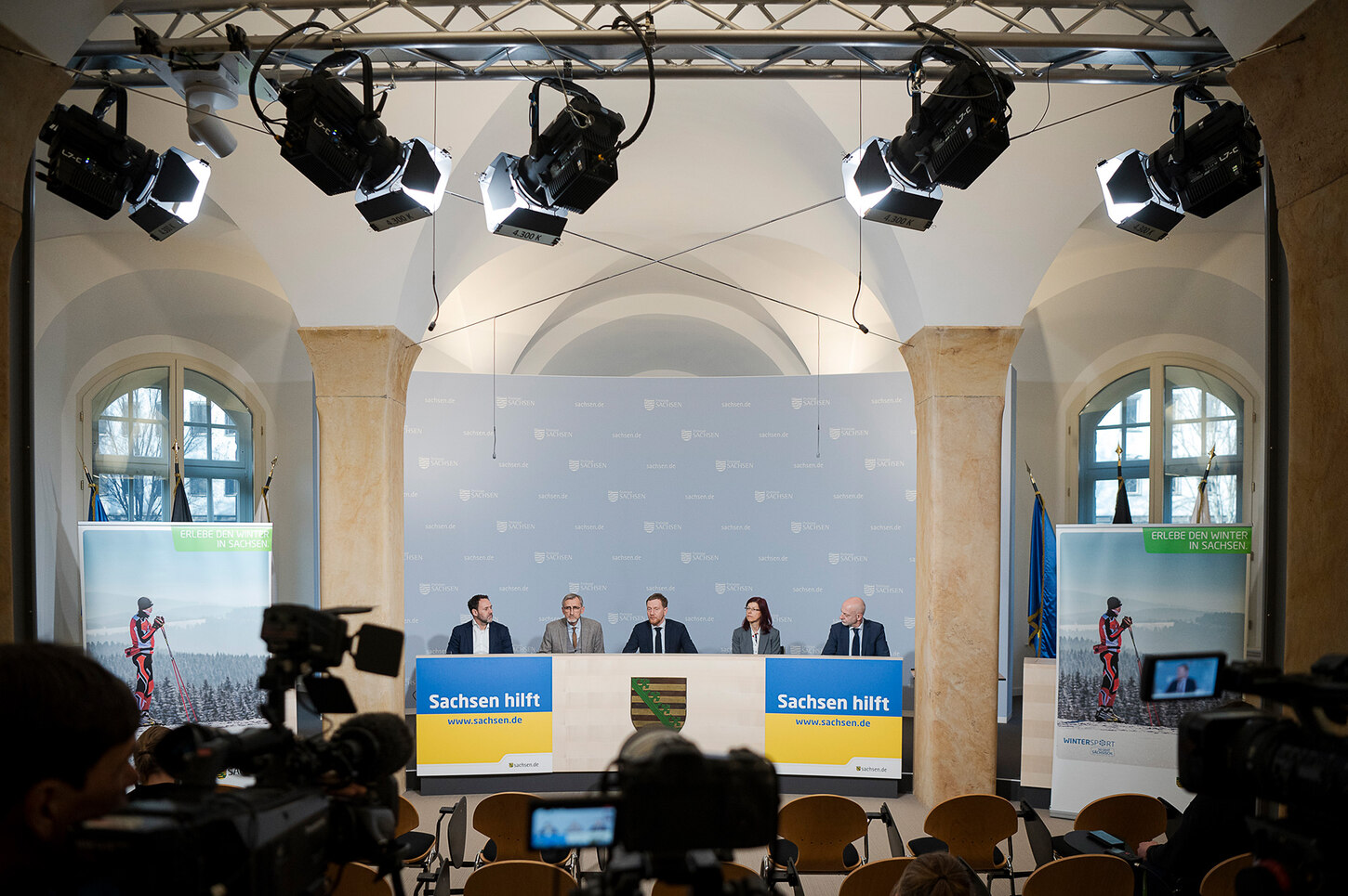 Männer sitzen auf einem Podium und berichten den im Saal versammelten Journalisten.