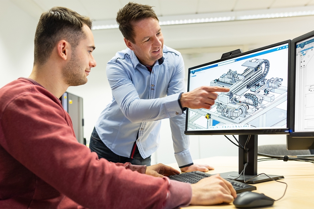 Ein junger Mann sitzt an einem Computerarbeitsplatz vor einem Monitor auf dem eine technische Grafik zu sehen ist. Ein zweiter Mann steht gebeugt neben dem Tisch und erklärt die technische Grafik.