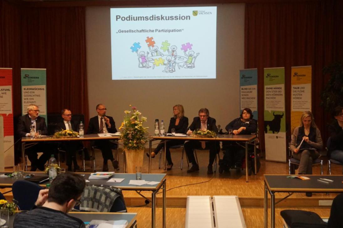 Experten der Podiumsdiskussion zur Offenen Tagung des Beauftragten sitzen an zwei Tischen und hören den Ausführungen des aktuellen Redners zu