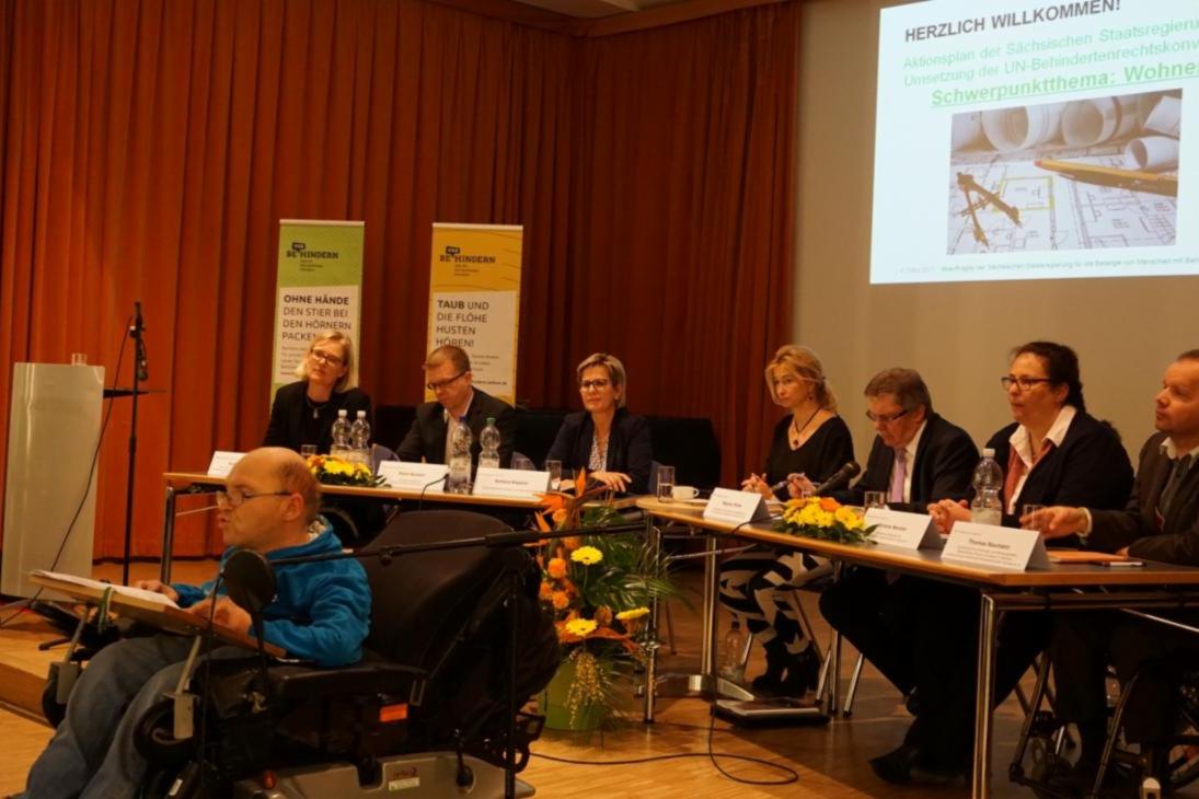 Experten der Podiumsdiskussion sitzen an zwei Tischen und hören den Ausführungen des aktuellen Redners zu, der vor ihnen in einem Rollstuhl sitzend ins Mikrophon spricht